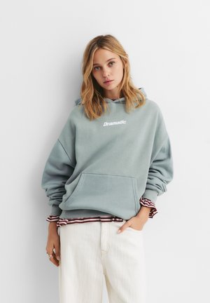 Jasnoniebieska, oversize bluza z kapturem z przednią kieszenią, z białym haftowanym napisem "Dramatic." Białe spodnie i w paski koszula pod spodem.