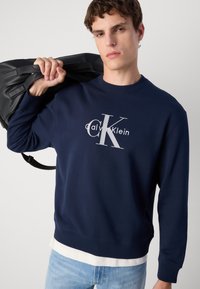 Sweatshirt azul-marinho com gola redonda e punhos canelados, adornado com um logotipo branco da Calvin Klein. Modelo segura uma bolsa preta.