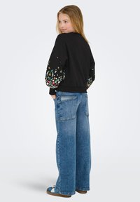 Svart sweatshirt med mönstrad, färgglad blommig brodyr på ärmarna. Kombineras med vida blå jeans och avslappnade skor.