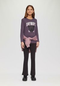 Lange mouw paarse shirt met een grafische print en tekst, gecombineerd met zwarte flared broeken en een licht paarse trui om de taille geknoopt. Zwarte schoenen.