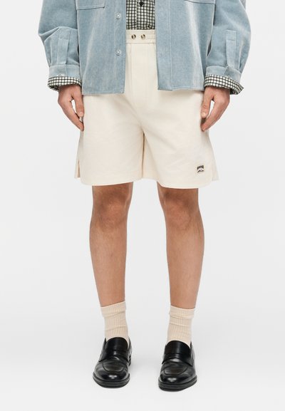 Homme portant un short crème avec des fentes latérales, une chemise bleue boutonnée, une chemise intérieure à carreaux, des chaussettes crème et des mocassins noirs, debout droit.