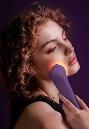 Purple facial device with an orange glowing tip held against the cheek, featuring a smooth texture and ergonomic design.