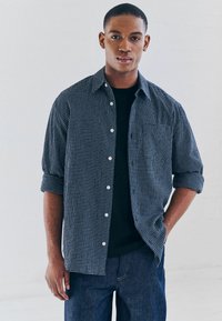 Camisa de cuadros azul y blanca de botones con las mangas remangadas, llevada sobre una camiseta negra, combinada con jeans de mezclilla oscura.
