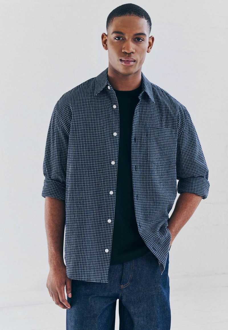 Camisa de cuadros azul y blanca de botones con las mangas remangadas, llevada sobre una camiseta negra, combinada con jeans de mezclilla oscura.