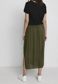 Jupe maxi plissée vert olive avec de hautes fentes latérales, associée à un haut noir à manches courtes et des baskets blanches.