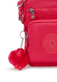 Röd nylon crossbody-väska med en texturerad yta, som har en framfickasdragkedja, en rund logotyp och en fluffig rosa apa nyckelring.