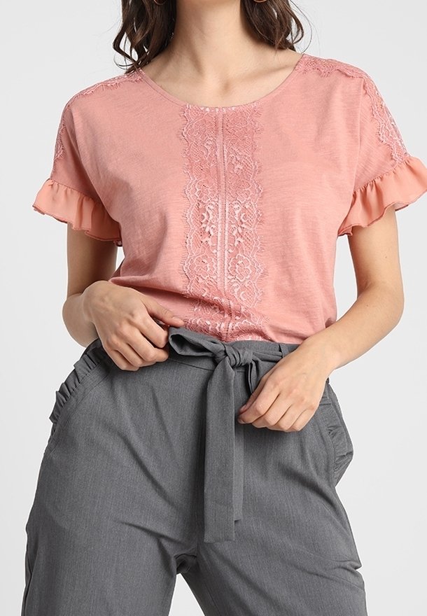 Kvinna iklädd en kortärmad topp i rosa med spetsdetaljer och volangkanter, samt högmidjade grå byxor som binds med ett tygbälte.