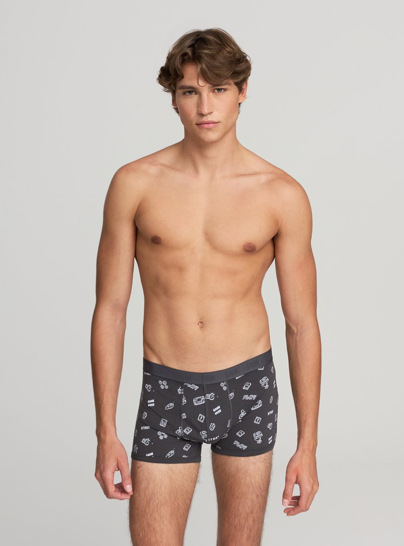 Młody mężczyzna stoi, ubrany w ciemne bokserki z białymi ikonami i tekstem związanymi z grami, na jednolitym jasnym tle.