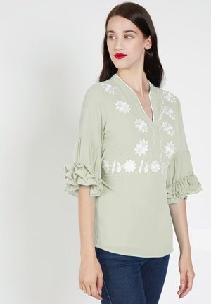 Blusa verde claro con bordado floral blanco, diseño de escote en V y mangas con volantes, combinada con jeans azul oscuro. Textura de tela suave.