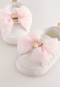 Witte babyschoentjes met een reliëf bloemmotief, voorzien van een grote roze doorschijnende strik en een metalen gouden gesp aan de voorkant. Zacht materiaal.