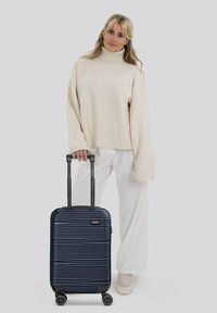 Valigia da viaggio blu navy con texture a coste orizzontali, resistente manico telescopico nero e quattro ruote, abbinata a un leggero maglione e pantaloni bianchi.