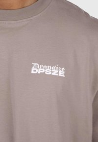 Graues T-Shirt mit kurzen Ärmeln aus Baumwolle. Auf der linken Brustseite befindet sich ein weißer Druck mit dem Logo "Droniżo DPSZE". Normale Passform, glatte Textur.