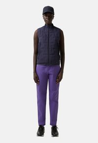 Marineblaue gesteppte Weste mit hohem Kragen, Reißverschluss, kombiniert mit lila Hose und schwarzen Turnschuhen. Ein flachkrempiges Cap rundet das Outfit ab.