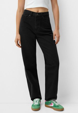 Jeans neri a vita alta con gambe dritte, caratterizzati da cinque tasche e chiusura con bottone. Abbinati a sneakers verdi con dettagli rosa.