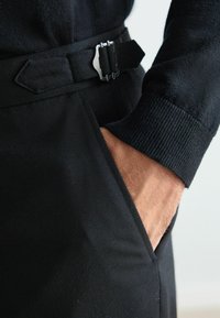 Schwarzer Strickpullover mit strukturiertem Finish, ausgestattet mit einem eng anliegenden Bündchen und einem schwarzen Gürtel mit einer silbernen Schnalle, wobei die Hand in einer Tasche positioniert ist.