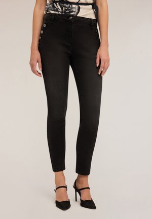 Femme portant un pantalon noir slim avec des boutons sur le côté et des chaussures noires à talons hauts avec bride à la cheville, debout devant un fond uni.