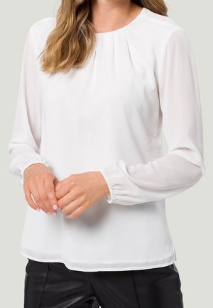 Femme portant un chemisier blanc à manches longues avec des poignets élastiques et des plis près de l'encolure, assorti à un pantalon noir.