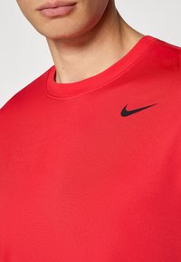 Nærbillede af en person iført en rød t-shirt med rund hals og et lille sort Nike swoosh-logo på venstre bryst.