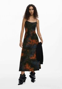 Rochie maxi fără mâneci cu un model rotitor în negru, verde și portocaliu, realizată dintr-un material moale. Însoțită de un strat exterior negru și cizme negre lucioase.