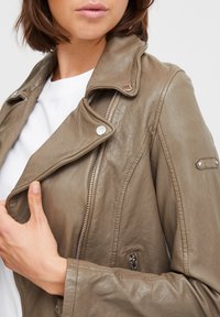 Khaki leather motorcycle jacket with zipped closure, snap buttons, and a textured finish. Features an angled collar and side pocket.