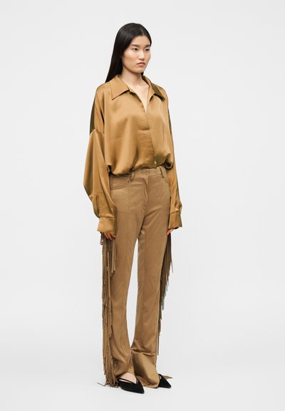 Μπλούζα σατέν χακί με άνετη εφαρμογή και φαρδιά μανίκια, σε συνδυασμό με χακί παντελόνι με μακριές φούντες κατά μήκος των πλευρών, συνδυασμένη με μαύρα παπούτσια.