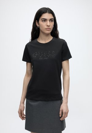 Kobieta o długich ciemnych włosach, ubrana w czarną koszulkę GUESS i szarą spódnicę, stojąca na jednolitym białym tle.