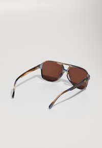 Ochelari de soare cu montură pătrată și lentile maronii, brațele din acetat cu model tartă, accent de logo auriu pe tamplă, având o montură transparentă de culoare gri-albastră.