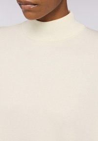 Maglione a collo alto di colore crema realizzato in morbido tessuto a maglia. Presenta un colletto alto e una texture liscia, con cuciture visibili minime.