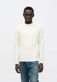 Μπλούζα πλεκτή σε κρέμ χρώμα με στρογγυλή λαιμόκοψη, ριμπ λεπτομέρειες και εφαρμοστή γραμμή, συνδυασμένη με μπλε τζιν. Ομαλή υφή.