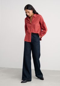 Roodbruine button-up blouse met pofmouwen, gecombineerd met wijde donkere spijkerbroeken en zwarte schoenen, tegen een neutrale achtergrond.