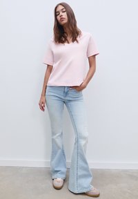 Helles rosa T-Shirt mit kurzen Ärmeln, kombiniert mit hellblauen Schlagjeans. Das Model trägt beige Sneaker mit einer strukturierten Oberfläche und hellen Sohlen.