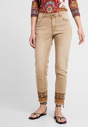 Beige broek in slim-fit met bloemenborduurwerk bij de pijpen, gedragen met zwarte sandaaltjes met bandjes en een top met lange mouwen en paisley-patroon.