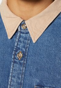 Colletto in corduroy beige su una camicia di denim blu con bottoni, indossata nella zona del collo e del petto superiore di una persona.