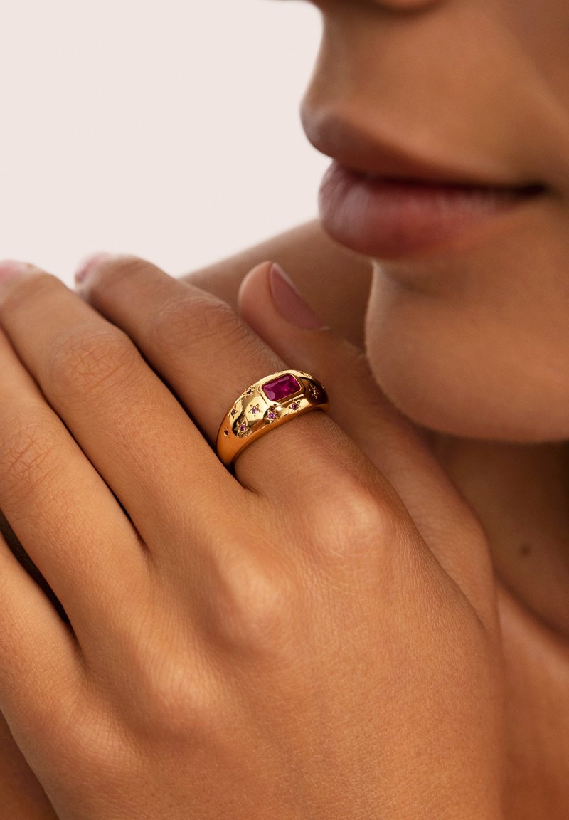 Goldring mit einem rechteckigen lila Stein, umgeben von kleineren Edelsteinen. Glatte Oberfläche mit subtilen Texturakzenten am Ring.