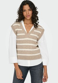 Väike beez kudumivest valgete triipudega, millel on V-kaelus ja ilma varrukateta disain, kihtide kaupa valge kaelusega särk. Näha on tume teksapüksid.