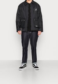 Chaqueta negra con cierre de cremallera, bolsillos de malla, logo bordado; combinada con jeans de mezclilla oscura y zapatillas negras con acentos blancos.