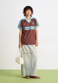 Kvinna i brun och turkos Adidas sporttröja, lång ljus denimkjol, hållande vit handväska, stående på grön golv mot vit vägg.