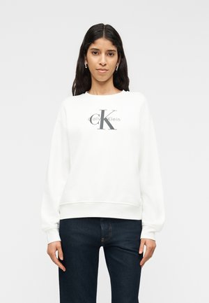 Γυναίκα με μακριά σκούρα μαλλιά, φορώντας λευκό φούτερ Calvin Klein και σκούρο μπλε τζιν, στέκεται μπροστά σε ένα απλό ανοιχτό φόντο.