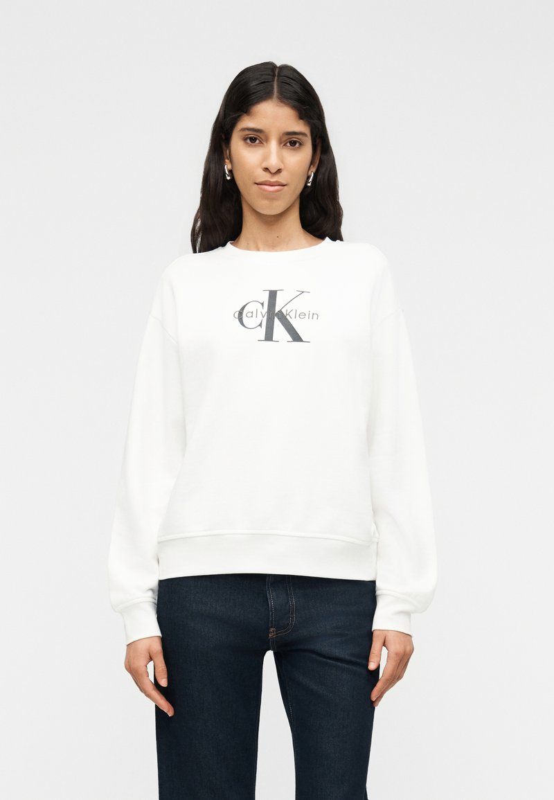 Γυναίκα με μακριά σκούρα μαλλιά, φορώντας λευκό φούτερ Calvin Klein και σκούρο μπλε τζιν, στέκεται μπροστά σε ένα απλό ανοιχτό φόντο.