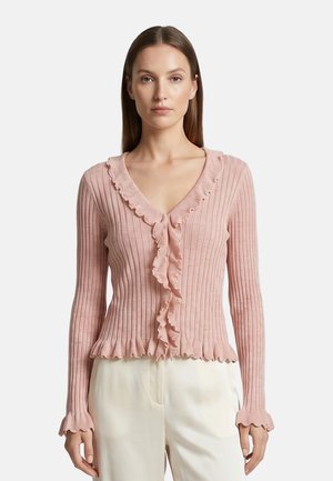 Vrouw draagt een lichtroze geribd vest met gerimpelde randen en crèmekleurige broek tegen een effen achtergrond.