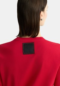 T-shirt rossa con una toppa nera quadrata sulla schiena, caratterizzata da cuciture arancioni a contrasto. Tessuto morbido e vestibilità rilassata.