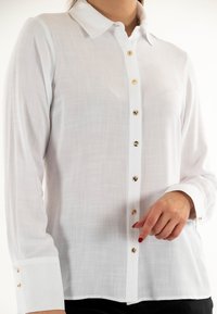 Camisa blanca de manga larga con cuello clásico, con botones dorados en la parte delantera y en los puños, hecha de una tela ligera y texturizada.