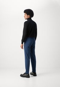 Fekete garbós pulóver, kék szabott nadrág és fekete bőrcipő. Simább textúrák, testre szabott sziluett. Minimalista design.