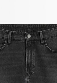 Section de la taille d'un jean en denim noir avec fermeture à bouton avant et passants pour ceinture, montrant une partie des poches avant.