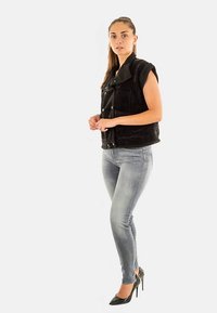 Svart ärmlös pälsväst med hög krage och tryckknappar, kombinerad med tajta ljusa jeans och svarta höga klackar, stående mot en vit bakgrund.