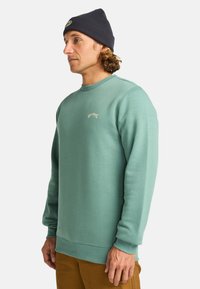 Felpa verde menta con polsini e orlo a costine, caratterizzata da un piccolo ricamo del logo sul petto. Abbinata a un berretto blu navy.