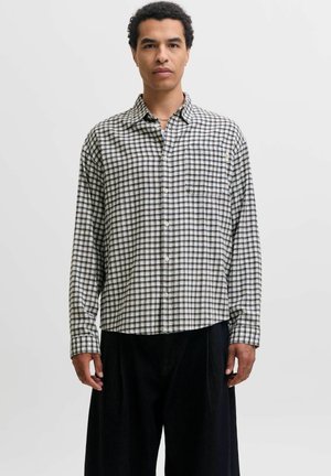 Homme portant une chemise à carreaux blanche et noire à manches longues et un pantalon noir, debout devant un fond clair uni.