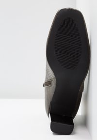 Semelle d'une bottine à bout carré avec une semelle extérieure en caoutchouc noire texturée et un petit talon carré, dessus en cuir brun clair à motif crocodile visible.