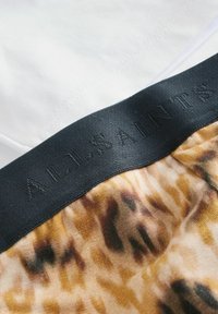 Kamelot tulostettu materiaali mustalla joustovyöllä, jossa on kohokuvioitu "A.L. Saints" -logo, kerrostettuna sileän valkoisen kankaan päälle.
