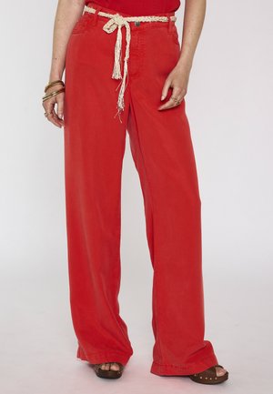 Personne portant un pantalon rouge à jambes larges avec une ceinture en corde tressée crème, des sandales marron, des bracelets dorés et un haut rouge, debout devant un fond uni.